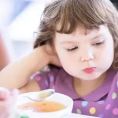 Mała dziewczynka z zaciekawieniem patrzy na łyżeczkę z zupą. Ciepłe kolory i widok jedzenia to coś, co naturalnie pobudza apetyt u dzieci.