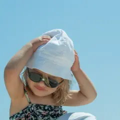 Dziecko w okularach przeciwsłonecznych i białej czapce UV dla dzieci, z rękami na głowie, na tle błękitnego nieba.