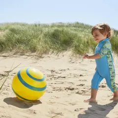 Chłopiec w błękitnym stroju kąpielowym dla niemowląt bawi się na plaży z żółtą piłką.