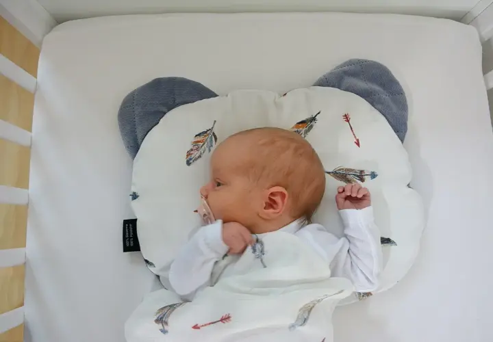 Nowy dzidziuś śpi na poduszce dla niemowlaka w kształcie misia, otulony kocykiem z piórkami.