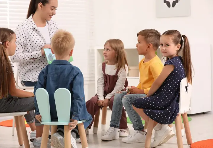 W przedszkolu nauczycielka czyta książkę grupie sześciorga dzieci. Wszyscy siedzą na krzesłach, słuchając uważnie.