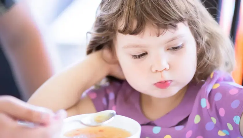Mała dziewczynka z zaciekawieniem patrzy na łyżeczkę z zupą. Ciepłe kolory i widok jedzenia to coś, co naturalnie pobudza apetyt u dzieci.