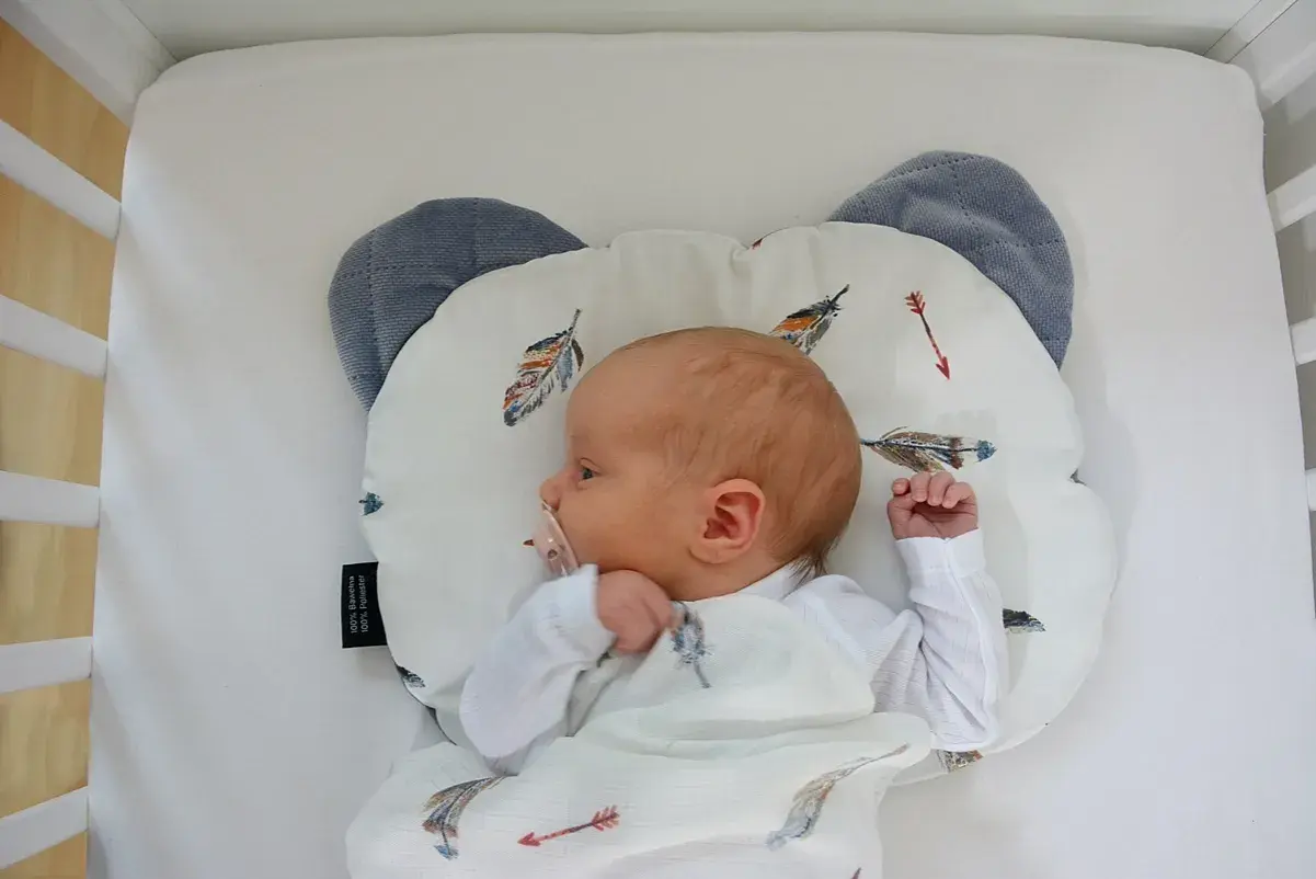 Nowy dzidziuś śpi na poduszce dla niemowlaka w kształcie misia, otulony kocykiem z piórkami.
