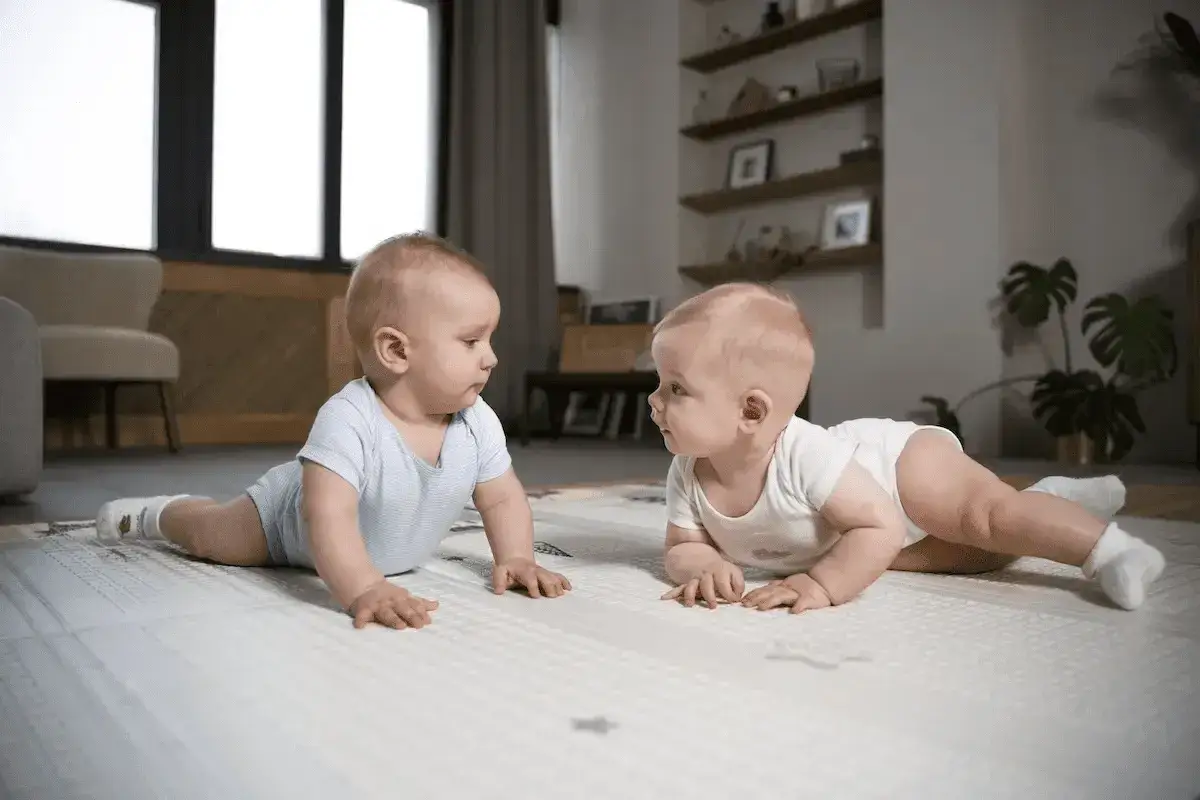 Dwa niemowlaki leżą na macie, badając się wzrokiem. Czy dziecko najpierw siada czy raczkuje?