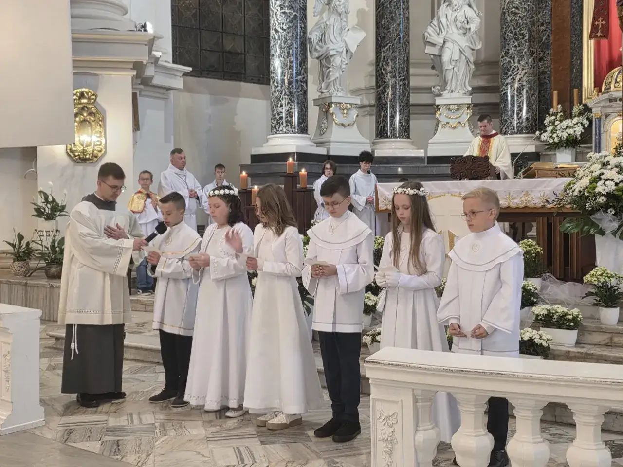 Dzieci w białych strojach komunijnych stoją w kościele. Czy rocznica komunii jest obowiązkowa? Duchowny przemawia.