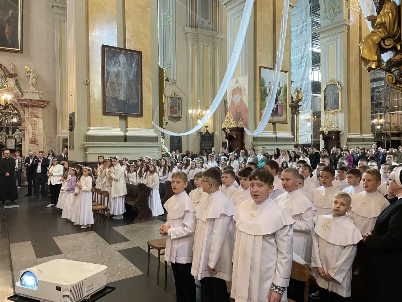 Dzieci w białych strojach komunijnych w kościele. Czy rocznica komunii jest obowiązkowa? Rodzice i goście obserwują uroczystość.